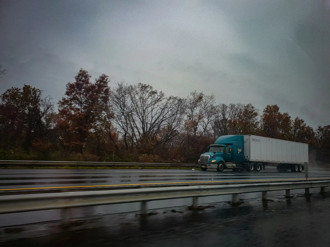 A green truck pulling a white trailer driving on a tree-and-fence-lined road, with blurring to indicate motion, connecting to the question, “What is the best GPS for truck drivers?”